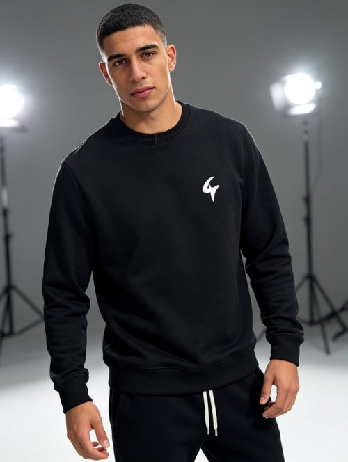 A man modeling a black crewneck sweatshirt with a small white logo on the left chest, paired with black track pants with white drawstrings, set against a gray studio background with softbox lights.