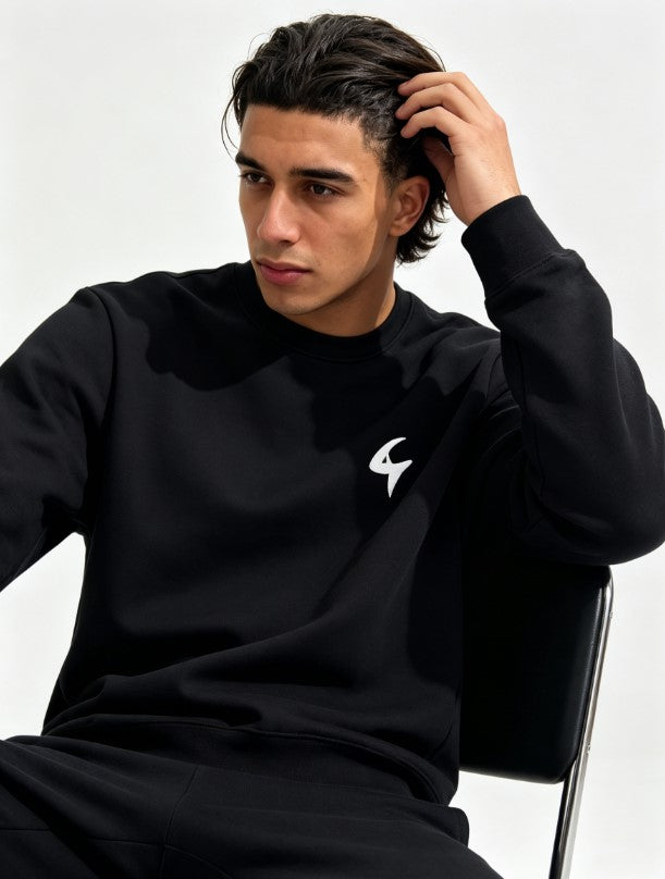 A man wearing a black crewneck sweatshirt with a small white stylized logo on the left chest, sitting against a plain light background.