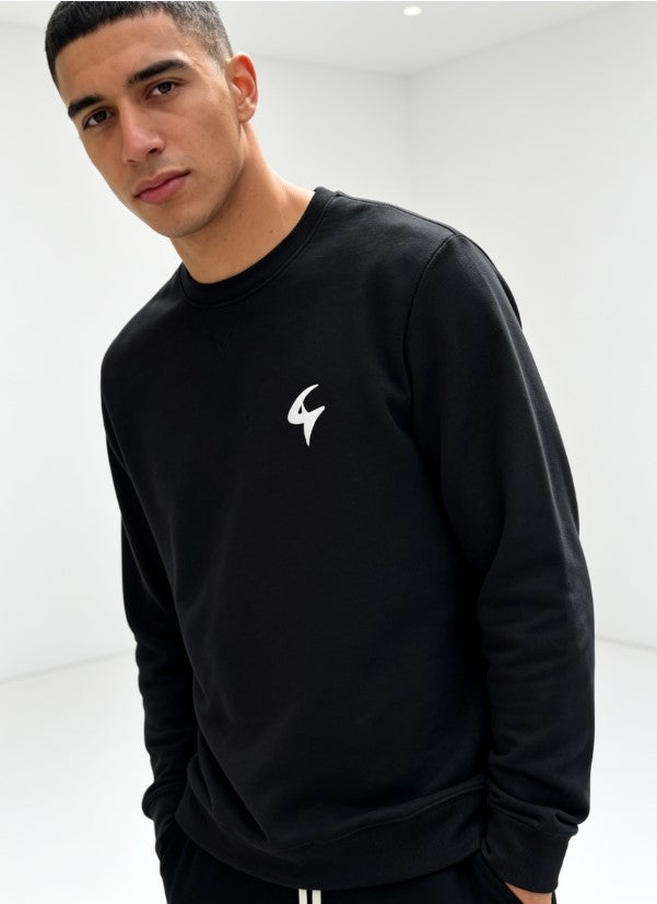 A man wearing a black crewneck sweatshirt with a small white stylized logo on the left chest, photographed in a bright white studio setting.