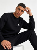 A man wearing a black crewneck sweatshirt and matching black sweatpants, sitting against a light gray studio background. The sweatshirt features a small white stylized logo on the left chest.