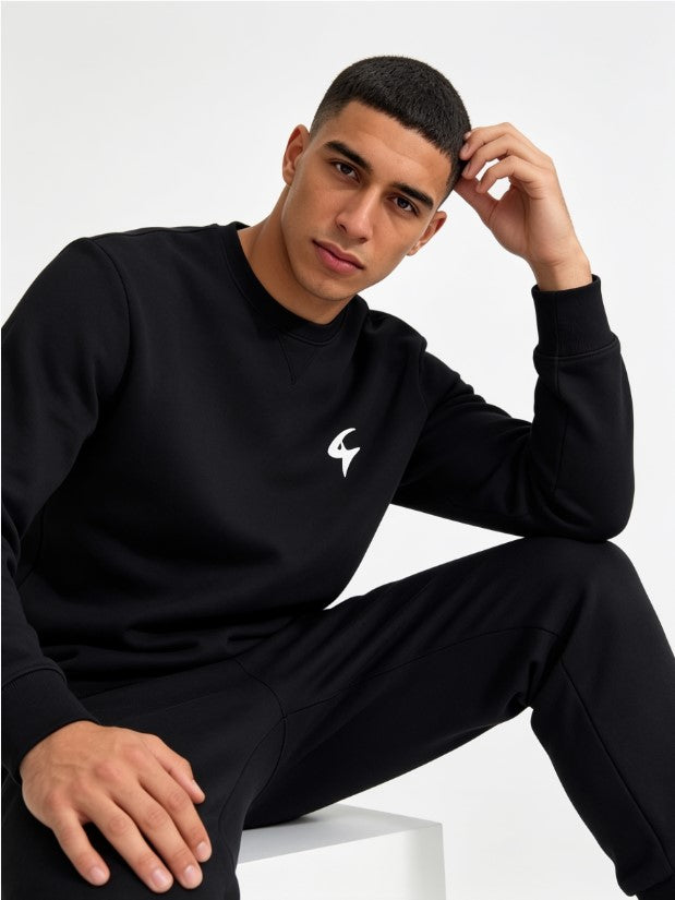A man wearing a black crewneck sweatshirt and matching black sweatpants, sitting against a light gray studio background. The sweatshirt features a small white stylized logo on the left chest.