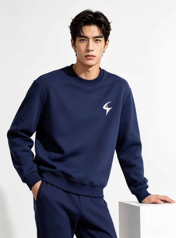 A man wearing a navy blue crewneck sweatshirt and matching sweatpants, featuring a small white stylized logo on the left chest, posed against a clean white studio background.