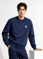 A man wearing a navy blue crewneck sweatshirt and matching sweatpants, featuring a small white stylized logo on the left chest, posed against a clean white studio background.