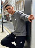 A man wearing a gray crewneck sweatshirt and black sweatpants with white drawstrings and logos, leaning against a modern pillar on a city street.