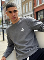 A man wearing a heather gray crewneck sweatshirt with a small white logo on the left chest, sitting outdoors on a white bench with a blurred street background.