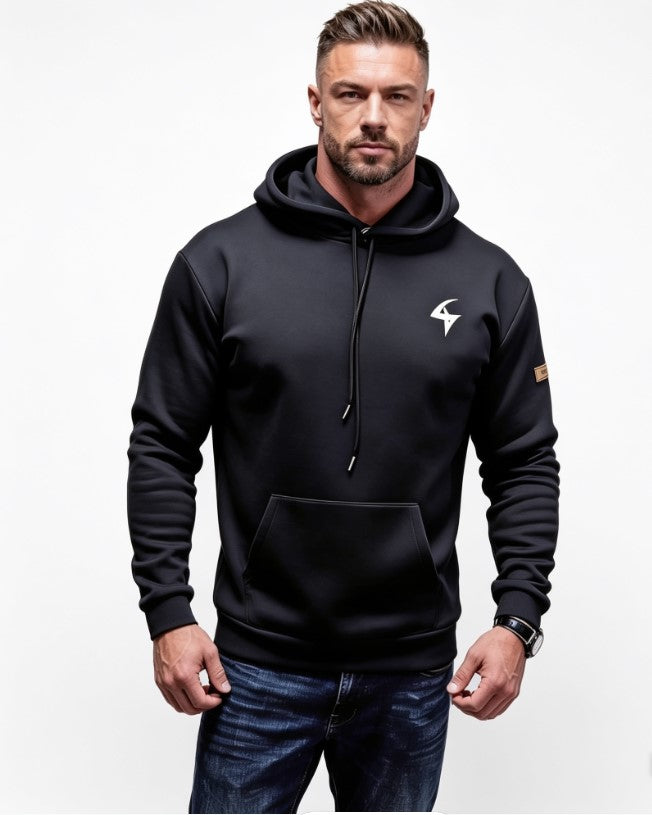 A man wearing a black pullover hoodie with a white stylized logo on the left chest and a small brown patch on the right sleeve, paired with dark blue jeans, against a plain white background.