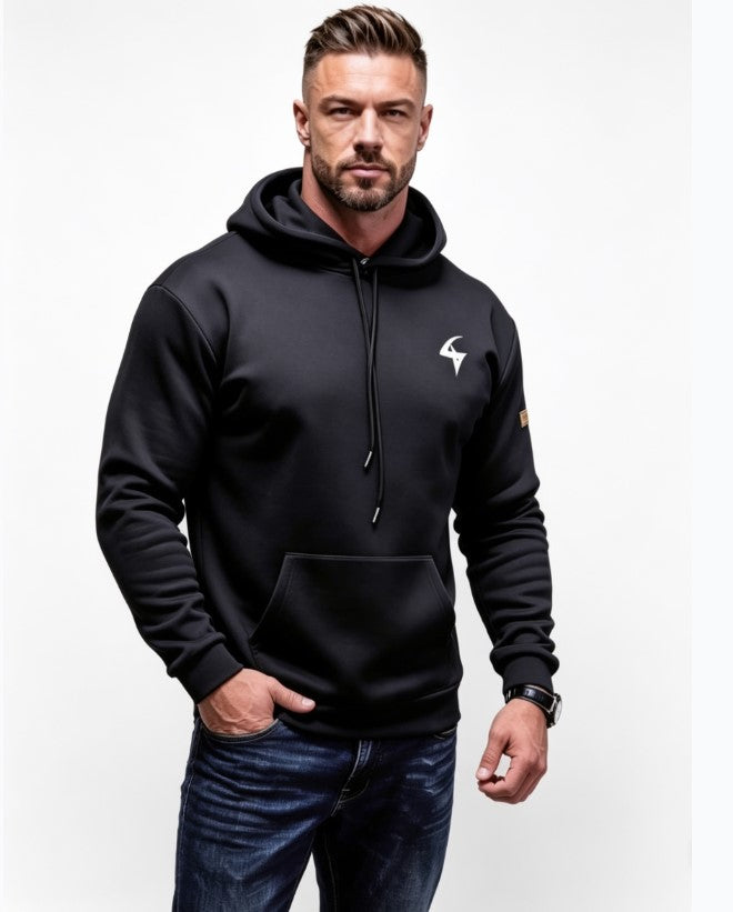 A man wearing a black pullover hoodie with a white graphic logo on the left chest and a small brown patch on the right sleeve, paired with dark blue jeans, against a plain white background.