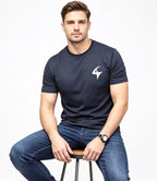 A man wearing a navy blue short-sleeve athletic t-shirt with a white stylized logo on the left chest, paired with blue jeans, sitting on a stool against a white background.