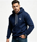 A man wearing a navy blue pullover hoodie with white drawstrings and a white stylized logo on the left chest, paired with blue jeans, photographed against a light gray studio background.