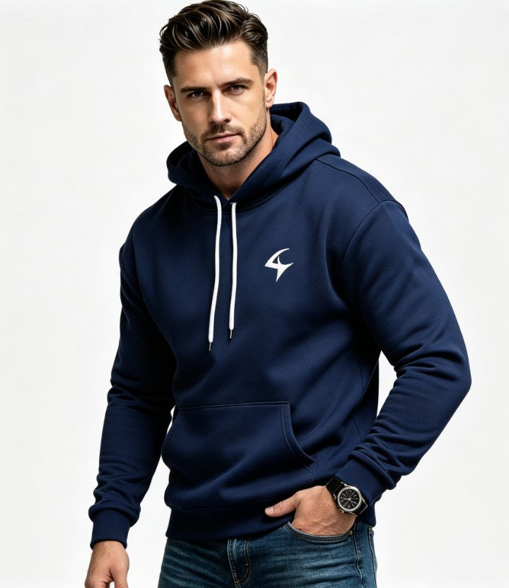 A man wearing a navy blue pullover hoodie with white drawstrings and a white stylized logo on the left chest, paired with blue jeans, photographed against a light gray studio background.