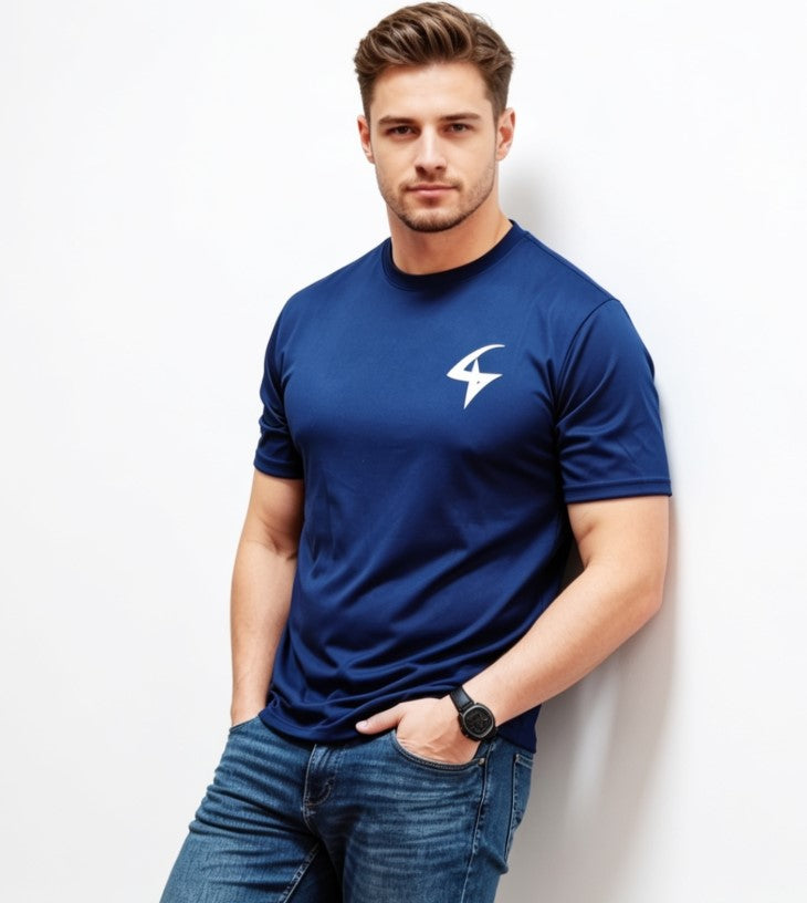 A man wearing a navy blue short-sleeve athletic t-shirt with a white logo on the left chest, paired with blue jeans, standing against a plain white wall.