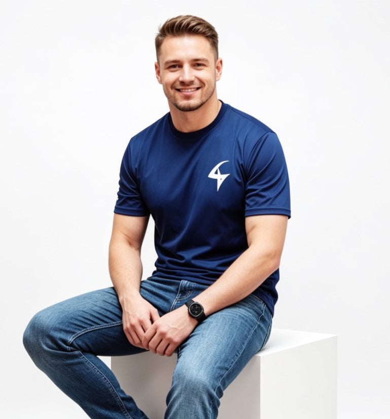 A smiling man wearing a navy blue athletic t-shirt with a white logo on the left chest, paired with blue jeans, sitting on a white block against a white studio background.