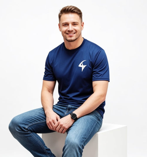A smiling man wearing a navy blue athletic t-shirt with a white logo on the left chest, paired with blue jeans, sitting on a white block against a white studio background.