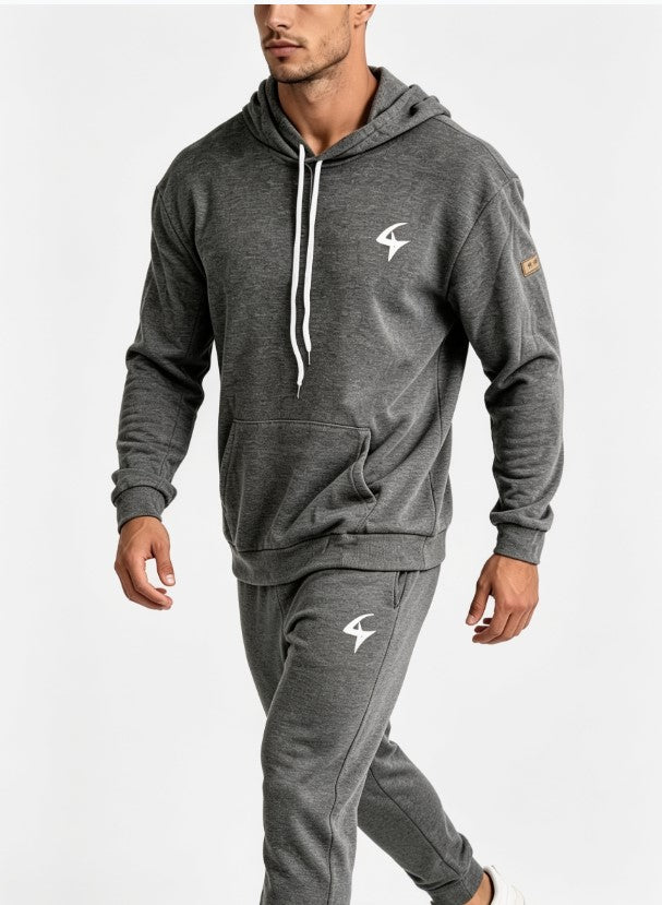 A man wearing a dark gray heathered hoodie and matching jogger pants with white drawstrings and white logo graphics, photographed against a clean white studio background.