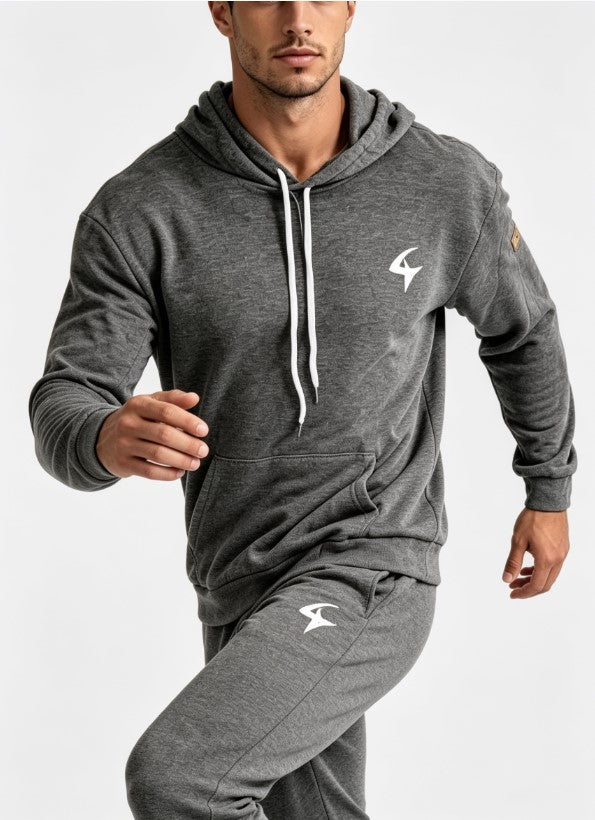 A man wearing a dark gray heathered athletic tracksuit with a matching hoodie and joggers, featuring a white logo on the chest and thigh, set against a plain light gray background.