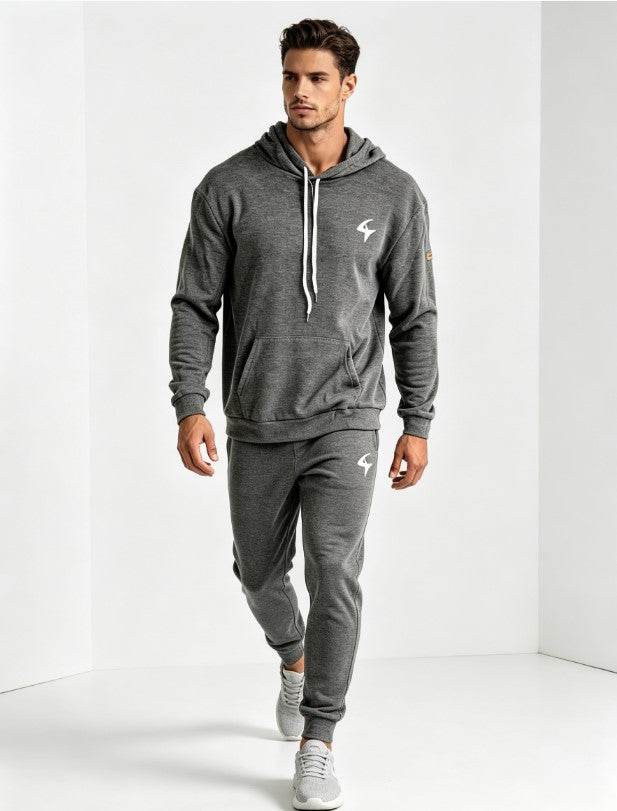 A man modeling a dark gray heathered athletic tracksuit, featuring a pullover hoodie with white drawstrings and a matching jogger pants set, both with small white logo graphics, set against a clean white studio background.