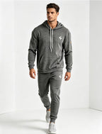 A man modeling a dark gray heathered athletic tracksuit, featuring a pullover hoodie with white drawstrings and a matching jogger pants set, both with small white logo graphics, set against a clean white studio background.