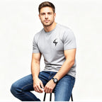 A man wearing a light gray short-sleeve t-shirt with a black lightning-bolt logo on the left chest, paired with blue jeans, sitting against a plain white background.