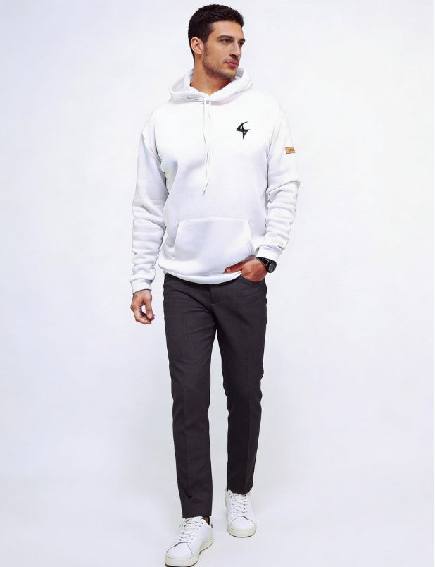 A man wearing a white pullover hoodie with a small black  logo on the left chest and a small brown patch on the right sleeve, paired with dark gray trousers and white sneakers, standing against a plain white studio background.