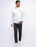 A man wearing a white pullover hoodie with a small black  logo on the left chest and a small brown patch on the right sleeve, paired with dark gray trousers and white sneakers, standing against a plain white studio background.